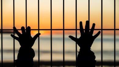 صورة ليدين لشخص يبدو في مكان مظلم وقد تعلقتا بقضبان حديدية تمنعهما عن الحرية