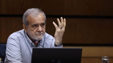 بزشكيان يرتدي قميصاً رمادي اللون، وساعة يد سوداء وخاتماً فضياً، شعره رمادي وبشرته حنطية، وأمامه مايكروفون ويحرك يده متحدثاً.