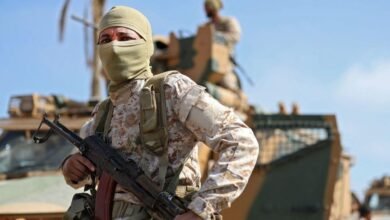 عنصر من قوات موالية للحكومة التي يترأسها دبيبة، يرتدي لباساً عسكرياً ويحمل سلاحاً بيديه ويغطي وجهه، وخلفه مركبة عسكرية.