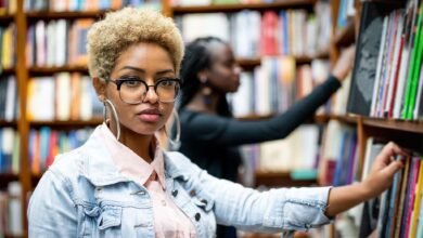 طالبة جامعية في مكتبة ترتدي سترة من الدنيم، تنظر نحو الكاميرا بينما تضع يدها على كتاب.