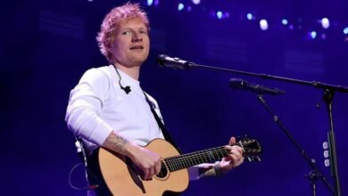 Ed Sheeran, in a long sleeved white T-shirt, holds a guitar and sings into a microphone. He is looking slightly off to one side.
