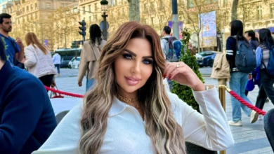سارة خليفة، ترتدي قميصا أبيض طويلاً، وذراعها اليسرى مطوية، ويدها تلمس شعرها الطويل، تجلس على جانب طريق داخل منطقة مطوقة بحبل أحمر، ويسير الناس خلفها على طريق تصطف على جانبه الآخر صفوف من المباني المشمسة.