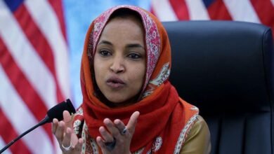تشير إلهان عمر بيدها أثناء حديثها. وهي جالسة على كرسي أسود وترتدي غطاء رأس برتقالي اللون بنقوش.