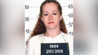 A woman with red hair stands in front of a sign that shows a person's hight, she wears a sign around her neck reading "TDOC 261368"