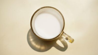 كوب زجاجي من الحليب يُرى من الأعلى، موضوع على خلفية باللون البيج ويلقي بظله عليها.