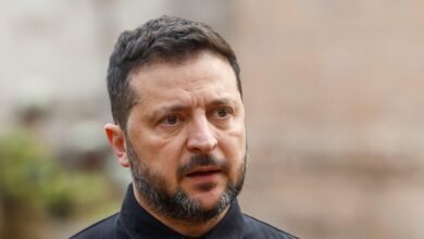 Ukranian President Volodymyr Zelensky wears a black shirt, his mouth is slightly open. The background behind him is blurred